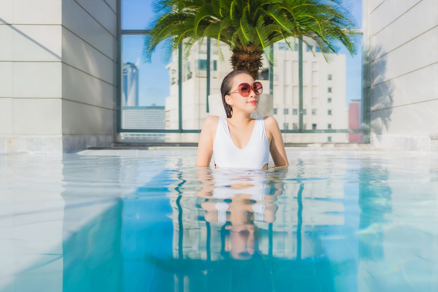 A woman in sunglasses and a white swimsuit stands in a swimming pool, with a palm tree and city buildings visible in the background, enjoying cool relief from heat waves.