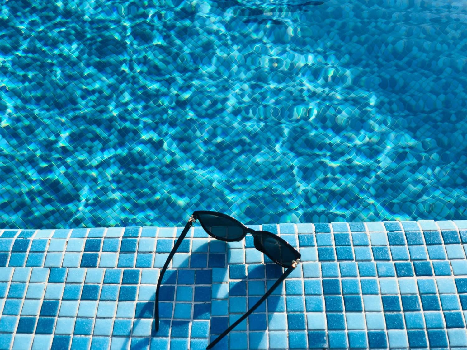 Black sunglasses rest on the edge of a tiled swimming pool, with sunlight reflecting on the water in the background—a reminder that regular pool filter maintenance keeps every swim sparkling clear.