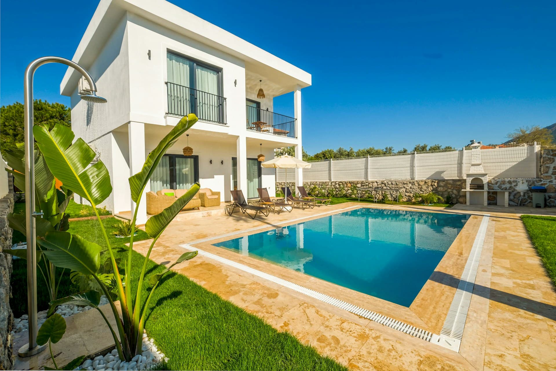 Modern two-story white house with balconies, a stone patio, sun loungers, and an outdoor pool surrounded by a manicured lawn and tropical plants—perfect for easy pool maintenance even during the cold months under a clear blue sky.