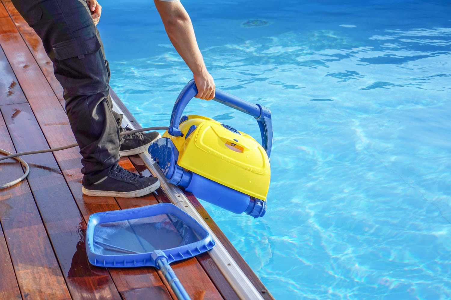 A person places a yellow and blue robotic pool cleaner into a swimming pool next to a wooden deck, with a pool skimmer net lying nearby—perfect for effortless Corpus Christi pool maintenance and beating the heat all season long.
