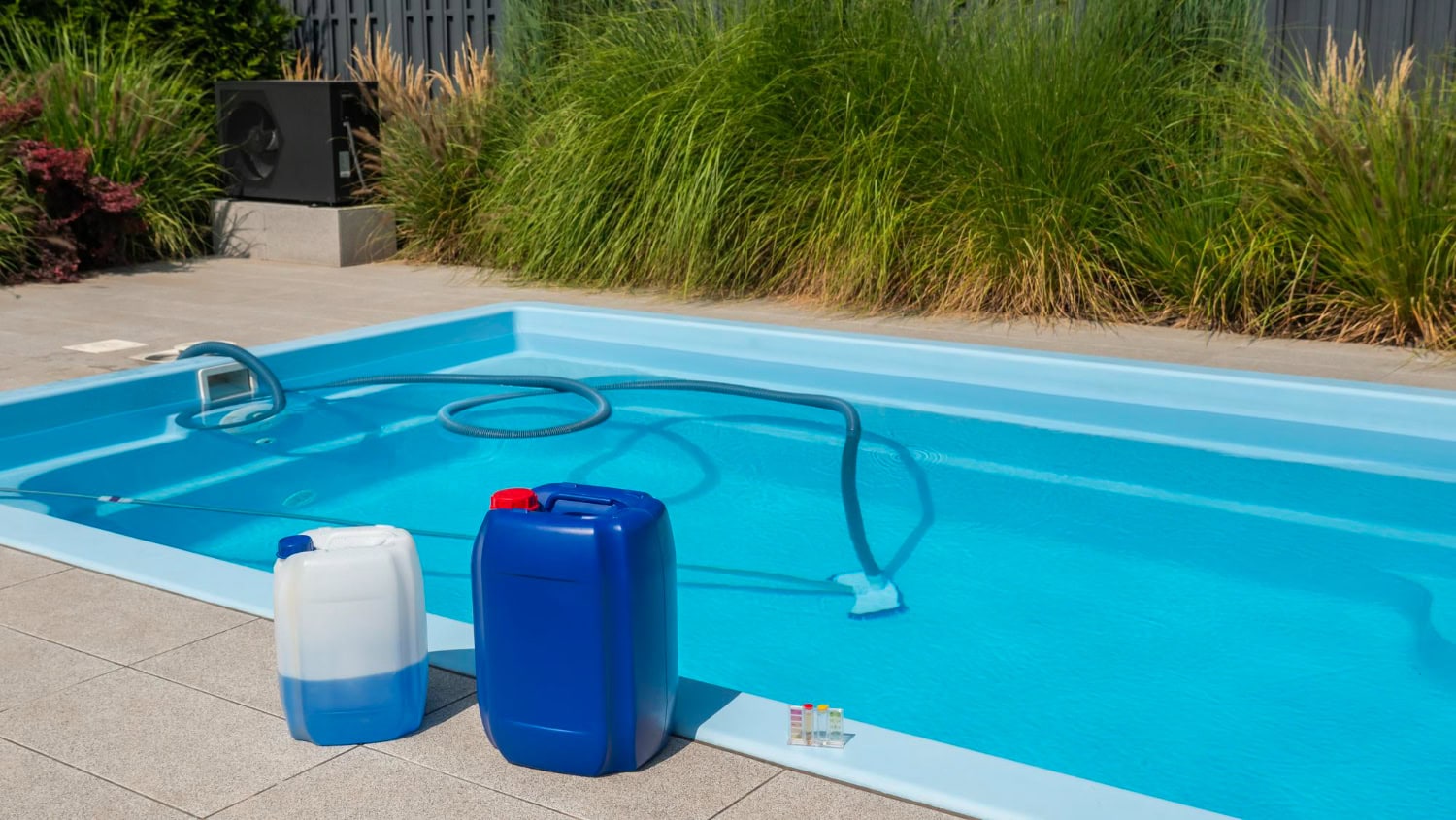 Two large chemical containers and a pool vacuum, essentials for pool maintenance, are beside an outdoor swimming pool, with plants and equipment in the background.